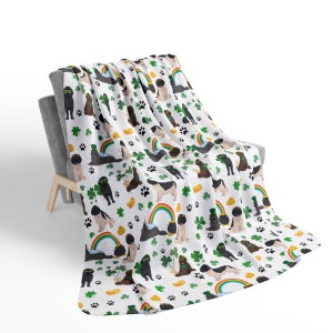 Peut inclure: Une couverture en polaire blanche avec un motif répété de chiens noirs portant des chapeaux verts et tenant des pièces d'or. Les chiens sont entourés de trèfles verts et d'arcs-en-ciel.