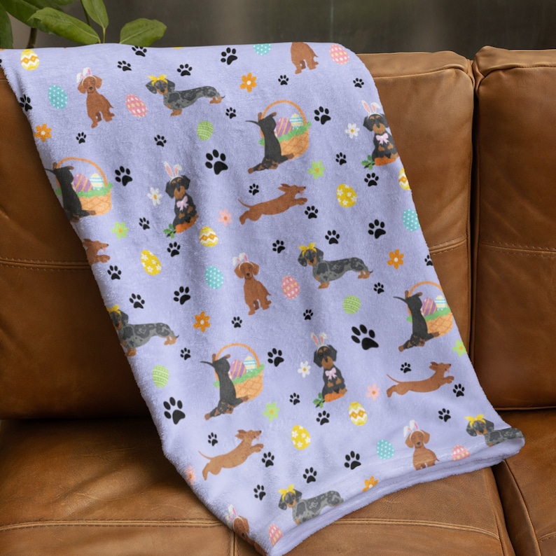 May include: A purple fleece throw blanket with a repeating pattern of dachshund dogs wearing bunny ears and holding Easter eggs. The pattern also includes paw prints, flowers, and Easter eggs.