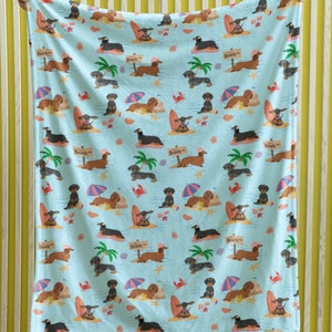 May include: A blue fleece blanket with a repeating pattern of dachshund dogs wearing sunglasses and surfing on surfboards. The dogs are surrounded by palm trees, seashells, and the words "Beach" on signs.