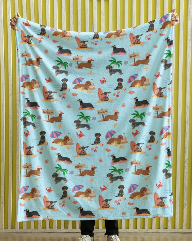 May include: A blue fleece blanket with a repeating pattern of dachshund dogs wearing sunglasses and surfing on surfboards. The dogs are surrounded by palm trees, seashells, and the words "Beach" on signs.