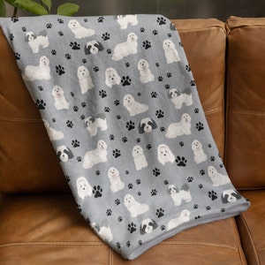 K&ouml;nnte beinhalten: Eine weiche graue Fleece-Decke mit einem sich wiederholenden Muster aus wei&szlig;en Havaneser-Hunden und schwarzen Pfotenabdr&uuml;cken.