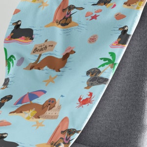 May include: A blue and white patterned throw blanket features a repeating pattern of dachshunds enjoying summer activities like surfing, building sandcastles, and relaxing on the beach. The text "Way to Beach" is featured on a sign.