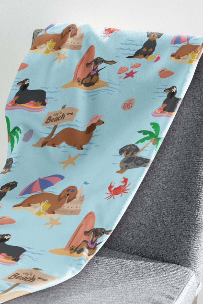 May include: A blue and white patterned throw blanket features a repeating pattern of dachshunds enjoying summer activities like surfing, building sandcastles, and relaxing on the beach. The text "Way to Beach" is featured on a sign.