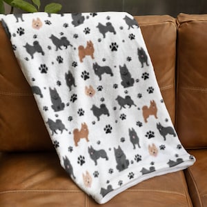 Puede incluir: Una manta blanca con un patrón repetido de huellas de patas negras e ilustraciones de perros en tonos marrón, beige y gris. La manta está sobre un sofá de cuero marrón.