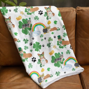May include: White fleece throw blanket with a repeating pattern of cartoon corgis wearing green shamrock hats and holding pots of gold. The corgis are surrounded by shamrocks, rainbows, and gold coins.