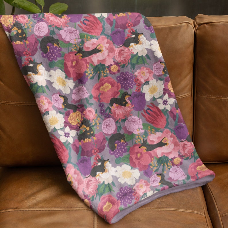 May include: A soft, purple and pink floral throw blanket with a repeating pattern of black dachshund dogs. The blanket is made of a soft, plush fabric and is perfect for cuddling up on the couch.