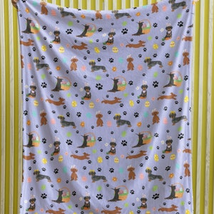 May include: A purple fleece throw blanket with a repeating pattern of dachshund dogs wearing bunny ears and holding Easter baskets. The dogs are brown and black with white accents. The background is a light purple with paw prints, flowers, and Easter eggs.