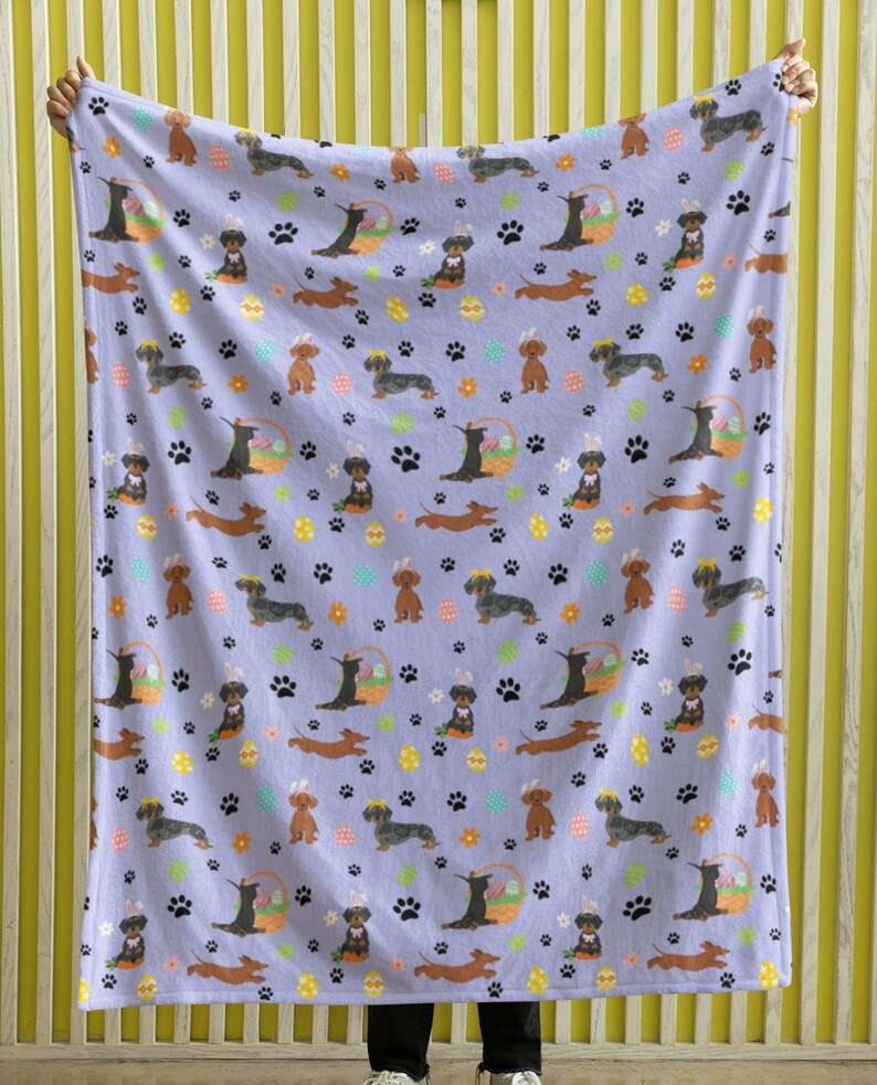 May include: A purple fleece throw blanket with a repeating pattern of dachshund dogs wearing bunny ears and holding Easter baskets. The dogs are brown and black with white accents. The background is a light purple with paw prints, flowers, and Easter eggs.