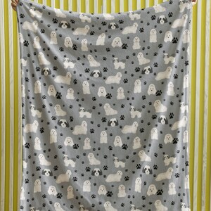 K&ouml;nnte beinhalten: Eine graue Fleece-Decke mit einem sich wiederholenden Muster aus wei&szlig;en Shih Tzu Hunden und schwarzen Pfotenabdr&uuml;cken.