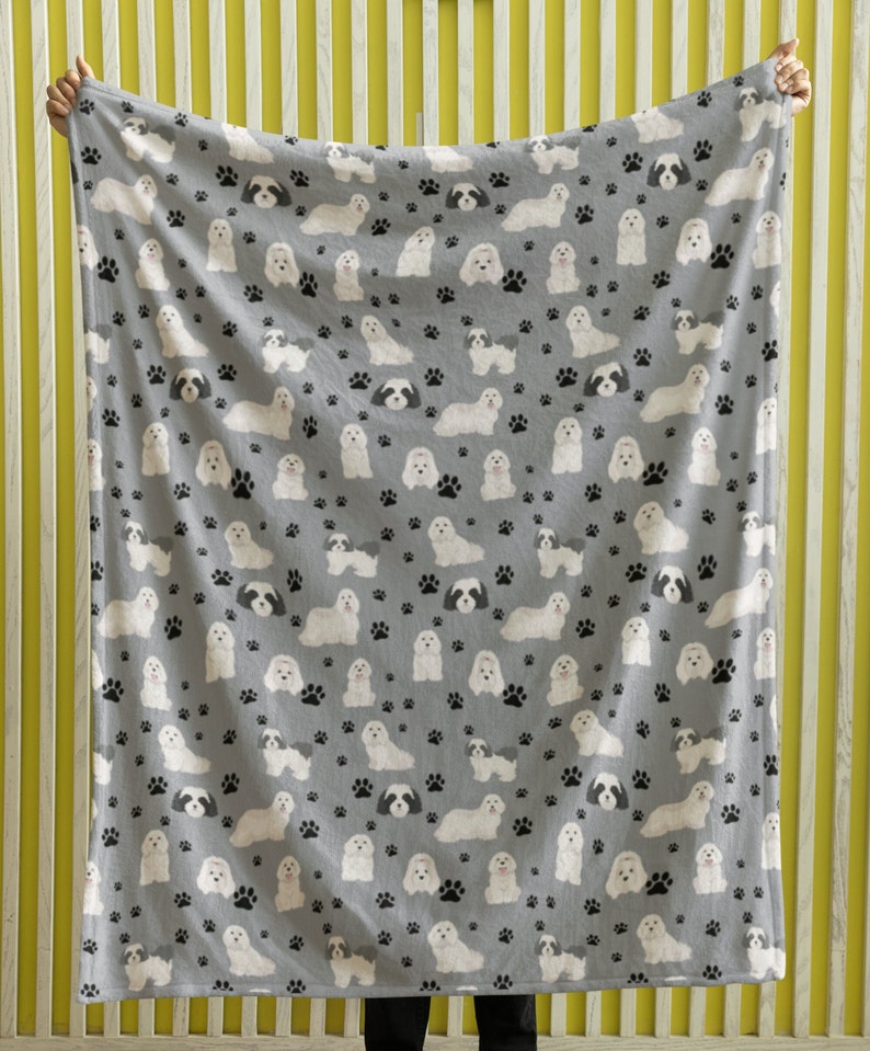 K&ouml;nnte beinhalten: Eine graue Fleece-Decke mit einem sich wiederholenden Muster aus wei&szlig;en Shih Tzu Hunden und schwarzen Pfotenabdr&uuml;cken.