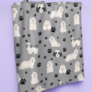 K&ouml;nnte beinhalten: Eine graue Fleece-Decke mit einem sich wiederholenden Muster aus wei&szlig;en Havaneser-Hunden und schwarzen Pfotenabdr&uuml;cken.