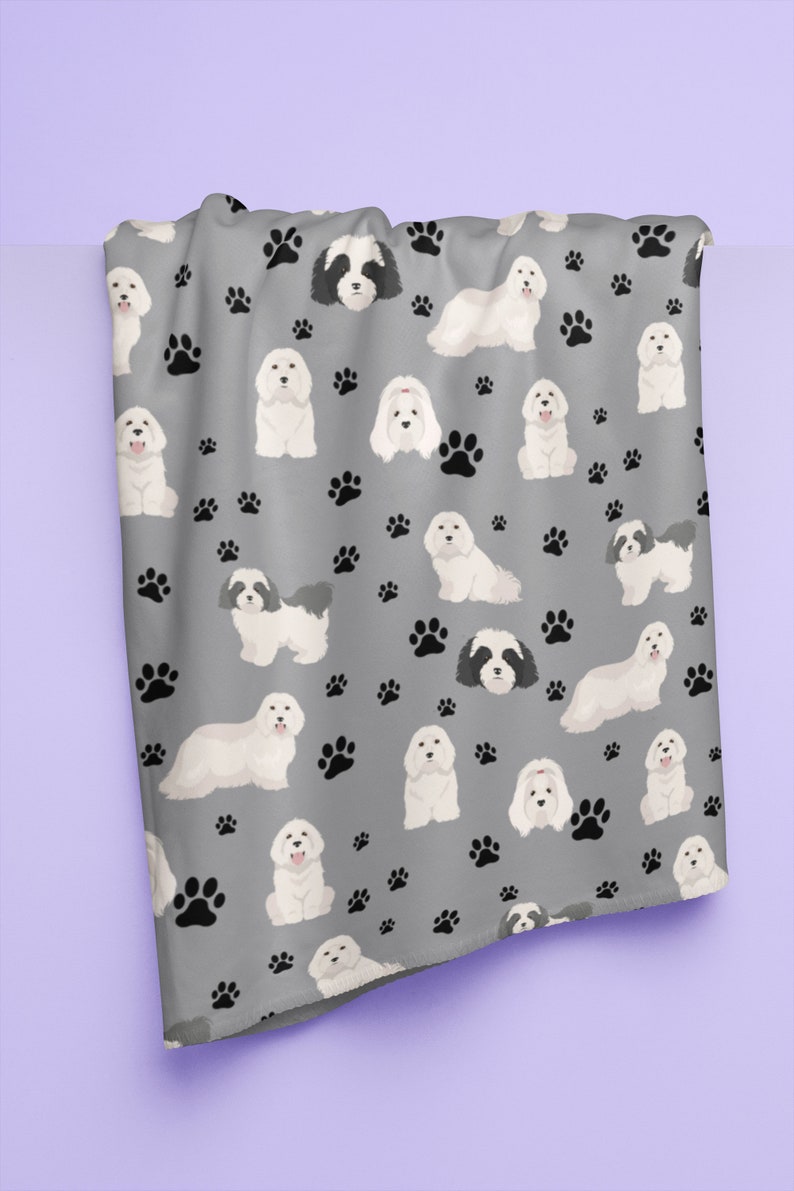 K&ouml;nnte beinhalten: Eine graue Fleece-Decke mit einem sich wiederholenden Muster aus wei&szlig;en Havaneser-Hunden und schwarzen Pfotenabdr&uuml;cken.