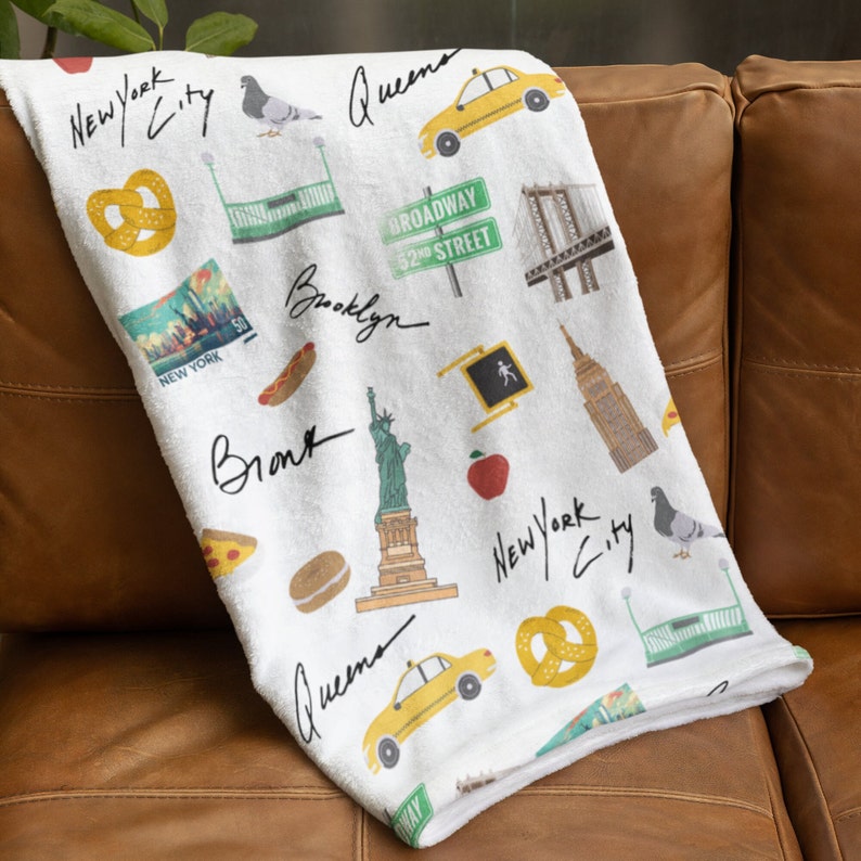 Puede incluir: Una manta blanca con tem&aacute;tica de la ciudad de Nueva York. Presenta ilustraciones de monumentos emblem&aacute;ticos, comida y transporte, incluyendo la Estatua de la Libertad, un taxi amarillo, un pretzel y una entrada de metro. Las palabras "New York City" est&aacute;n escritas en letra negra a trav&eacute;s de la manta.