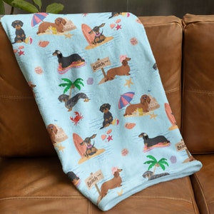 May include: A blue and white fleece throw blanket with a repeating pattern of dachshunds on the beach. The dachshunds are wearing sunglasses and holding surfboards. The text "Way to the Beach" is repeated on the blanket.