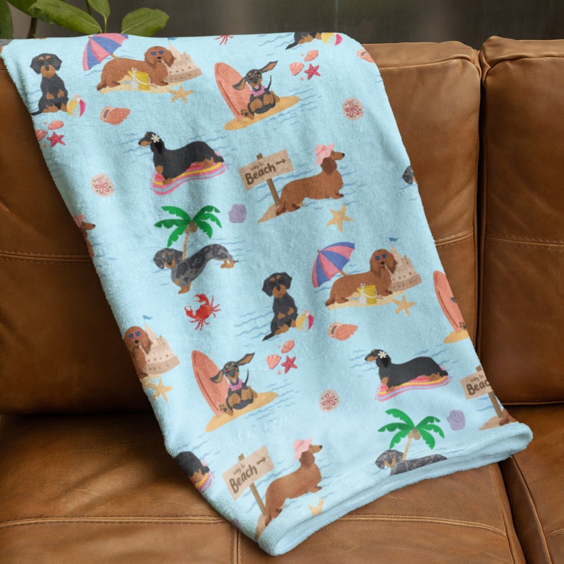 May include: A blue and white fleece throw blanket with a repeating pattern of dachshunds on the beach. The dachshunds are wearing sunglasses and holding surfboards. The text "Way to the Beach" is repeated on the blanket.