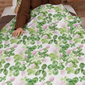 May include: White blanket with a repeating pattern of green clover leaves and pink flowers. The blanket is folded and a person's hand is reaching out from under the blanket.