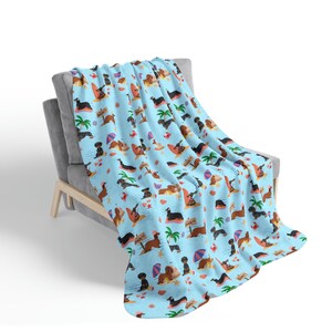 May include: A blue and white throw blanket with a pattern of dachshunds on a beach. The dachshunds are wearing sunglasses and holding surfboards. The blanket is on a gray couch.