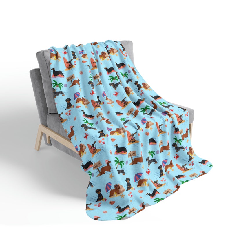 May include: A blue and white throw blanket with a pattern of dachshunds on a beach. The dachshunds are wearing sunglasses and holding surfboards. The blanket is on a gray couch.