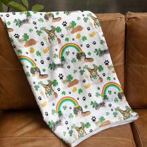 Puede incluir: Manta de forro polar blanca con un patrón repetido de corgis con sombreros verdes y sosteniendo tréboles. Los corgis están rodeados de arcoíris, tréboles y monedas de oro.