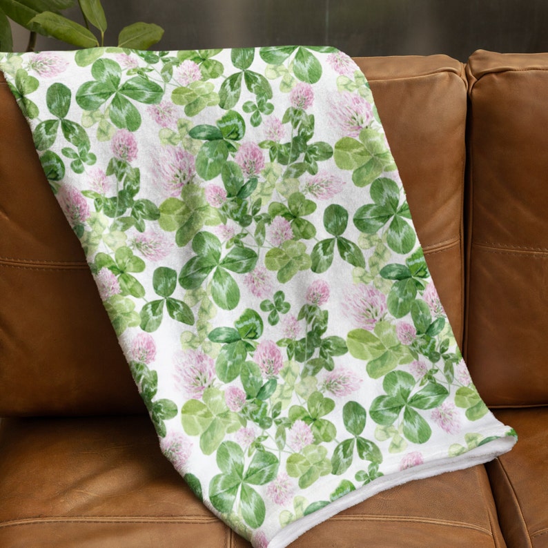 May include: A white throw blanket with a green clover and pink flower pattern. The blanket is folded and draped over a brown leather couch.