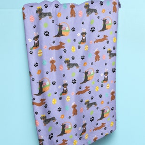 May include: A purple fleece blanket with a repeating pattern of dachshund dogs wearing bunny ears and holding Easter eggs. The dogs are brown and black with yellow and white accents. The background is a light purple with paw prints, flowers, and Easter eggs.