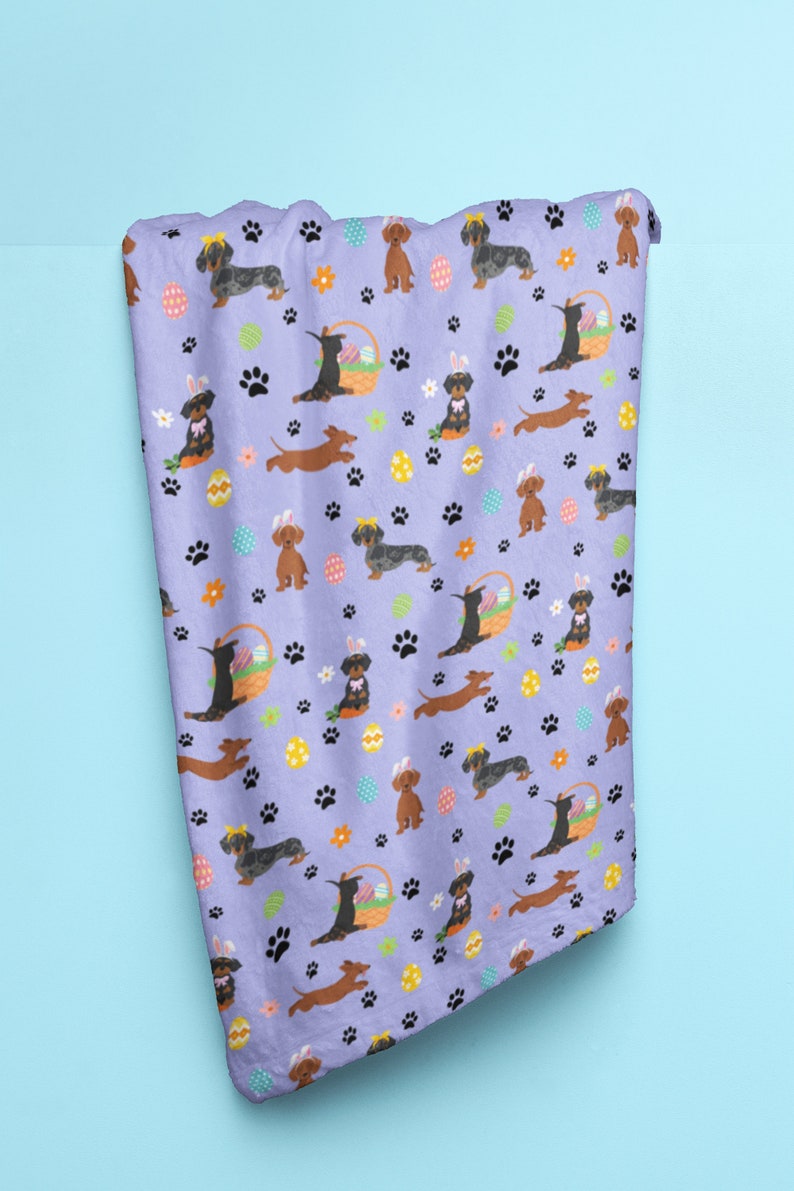 May include: A purple fleece blanket with a repeating pattern of dachshund dogs wearing bunny ears and holding Easter eggs. The dogs are brown and black with yellow and white accents. The background is a light purple with paw prints, flowers, and Easter eggs.