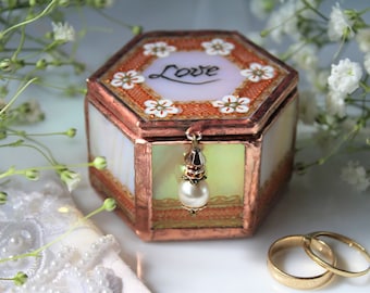 May include: A hexagonal glass ring box with a copper frame and a white lid with the word "Love" written in cursive. The lid is decorated with small white flowers and an orange pattern. A small pearl hangs from the lid.