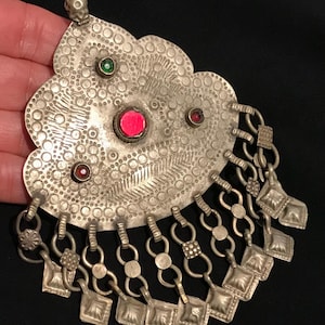 May include: Silver-toned pendant with intricate dot and line patterns, adorned with red and green gemstones. The pendant has a floral-like shape with dangling chain and square charms, creating a vintage aesthetic.