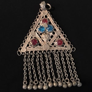 Puede incluir: Un colgante triangular de plata con piedras de vidrio rojo y azul y una franja de cadenas de plata con pequeñas bolas en los extremos.