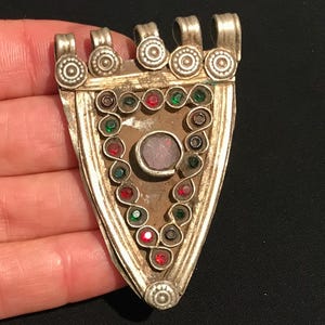 May include: Antique silver pendant with a triangular design. The pendant features a central pink stone, surrounded by a border of red and green gemstones. The top has decorative loops and circular accents. A vintage jewelry piece.