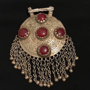 Könnte beinhalten: Silberfarbener Anhänger mit fünf roten Edelsteinen in einem kreisförmigen Design. Der Anhänger weist aufwendige geätzte Muster auf und ist mit herabhängenden Ketten und kleinen glockenförmigen Charms verziert. Der Hintergrund ist schwarz.