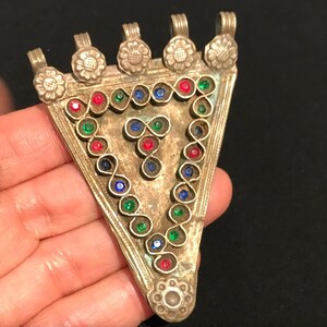 May include: Vintage silver pendant, triangular in shape, with a floral design and colourful gemstones. The pendant is decorated with red, green, and blue stones. The pendant is held in a hand.