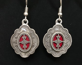 Puede incluir: Pendientes de plata con un diseño ovalado rojo y plateado. El óvalo tiene un patrón de cruz en el interior.