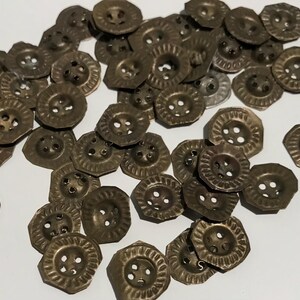 May include: A collection of antique brass-coloured buttons. The buttons have a decorative, octagonal shape with a textured rim and four holes for sewing. The buttons are scattered on a white surface.