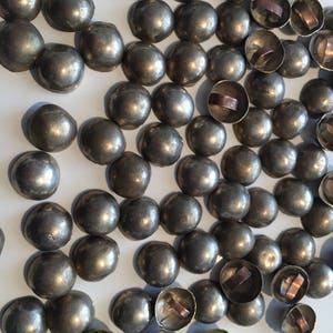 May include: A large group of silver-toned metal dome-shaped buttons with copper rings on the back. The buttons are arranged in a random pattern on a white surface.