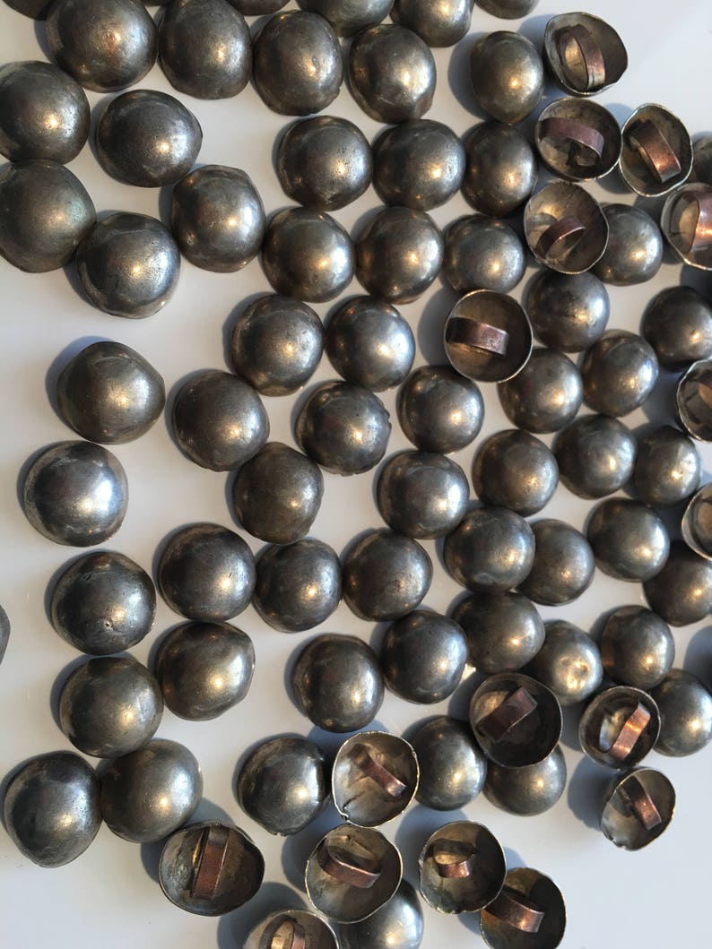 May include: A large group of silver-toned metal dome-shaped buttons with copper rings on the back. The buttons are arranged in a random pattern on a white surface.