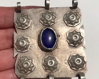 May include: A silver-toned pendant with a central oval blue stone and eight surrounding floral-shaped embellishments. The pendant has three loops for hanging and a slightly distressed appearance, suggesting an antique or vintage piece.