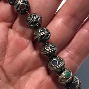 May include: A close-up of a silver beaded necklace with colorful stones. The beads are round and have a decorative design. The necklace is made of metal and has a vintage look.