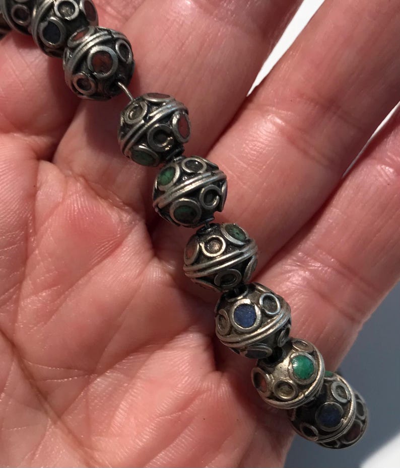 May include: A close-up of a silver beaded necklace with colorful stones. The beads are round and have a decorative design. The necklace is made of metal and has a vintage look.