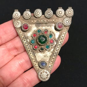 Puede incluir: Colgante triangular plateado antiguo con detalles intrincados. El colgante presenta un grupo central de piedras preciosas verdes, rojas y turquesas, rodeado por un borde decorativo. Se engastan piedras preciosas rojas y azules adicionales en el diseño. La parte superior tiene una fila de adornos circulares.