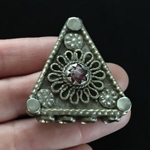 May include: A triangular silver pendant with a red stone in the center, surrounded by a decorative silver design. The pendant has a textured surface and is adorned with small silver beads.