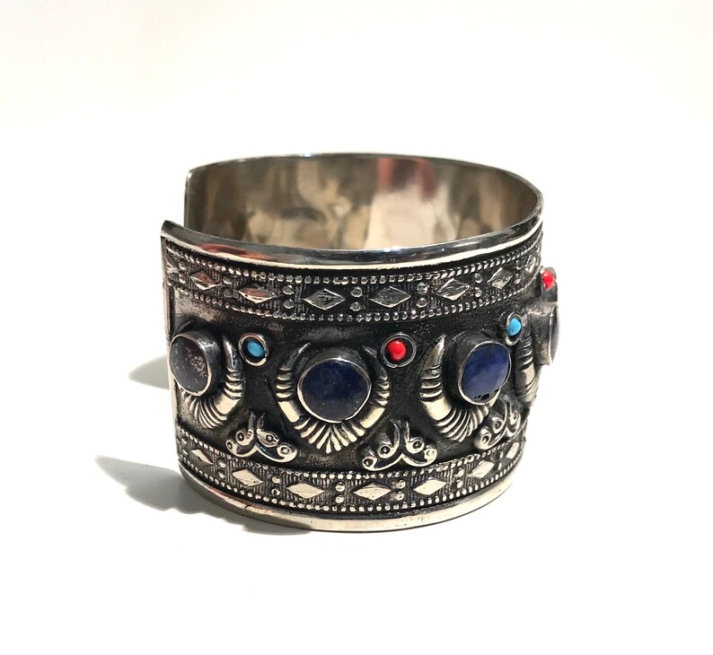 May include: Silver cuff bracelet with intricate detailing and blue and red gemstones.