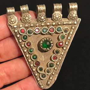 May include: A silver-coloured triangular pendant with red, green, and clear gemstones. The pendant has a central green stone and is adorned with decorative floral elements. The top features five loops for hanging.