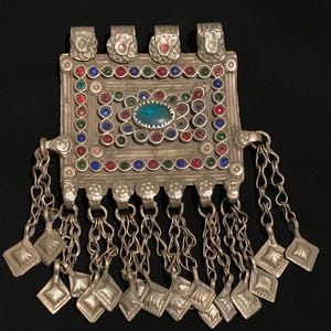 Könnte beinhalten: Silberner Anhänger mit rechteckigem Design, verziert mit farbigen Edelsteinen und einem zentralen ovalen blauen Stein. Der Anhänger verfügt über dekorative Metallarbeiten und baumelnde Ketten mit quadratischen Anhängern.