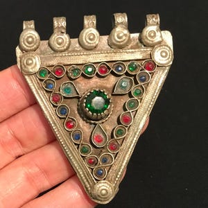 Puede incluir: Colgante triangular plateado con una gema verde central y una serie de gemas de colores. El colgante presenta piedras rojas, verdes y azules, y tiene cinco bucles en la parte superior para colgar. El diseño es intrincado y detallado.