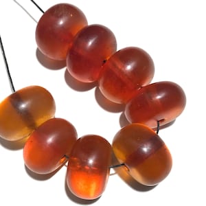 Könnte beinhalten: Eine Kette aus fassförmigen, bernsteinfarbenen Perlen. Die Perlen sind auf einer schwarzen Schnur aufgefädelt und haben ein durchscheinendes Aussehen, mit Farbvariationen von hellorange bis tiefrot. Die Perlen sind glatt und poliert.