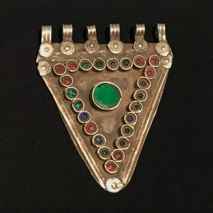 Puede incluir: Colgante triangular plateado con una gema verde central y un borde de gemas de colores. El colgante tiene una serie de bucles en la parte superior para colgar. Las gemas son rojas, verdes y azules.