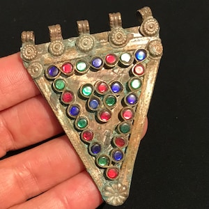 May include: Vintage triangular pendant with a tarnished gold finish. The pendant is decorated with colourful, round glass stones in red, green, and blue. The top has four decorative loops.
