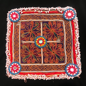 Könnte beinhalten: Ein quadratisches, lila und orangefarbenes, besticktes Stoffkissen mit einem weißen Perlenrand. Das Kissen hat ein geometrisches Muster mit vier floralen Motiven in der Mitte. Das Kissen ist mit bunten Perlen verziert.