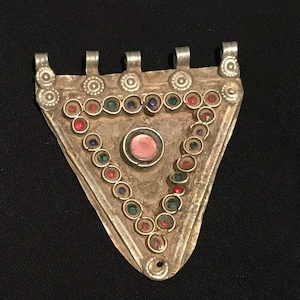 Puede incluir: Colgante triangular plateado con una piedra rosa central y un borde de gemas de colores. El colgante tiene bucles en la parte superior para colgar, probablemente una joya.